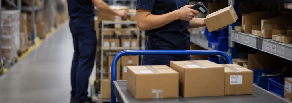 Operaria escaneando un paquete con lector de códigos de barras en un almacén, junto a estanterías con cajas y un sistema digital de gestión de pedidos.