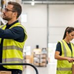Preparación de pedidos almacén: métodos y consejos Operarios preparando pedidos en un almacén moderno: uno realiza picking con lector de códigos de barras y otro embala un paquete en la zona de expedición.